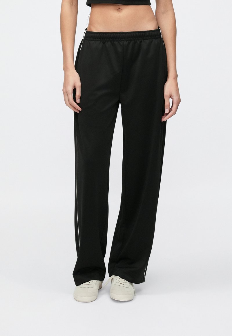 Žena v čiernych širokých teplákoch s bielymi bočnými pruhmi a bielobielymi teniskami, stojaca na jednoduchom bielom pozadí.