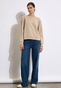 Misdina beige a maniche lunghe con decorazioni a borchie disposte a V, abbinata a jeans blu a gamba larga e sneakers bianche. Sfondo semplice.