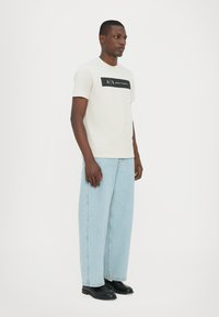 Lys grå T-skjorte med svart "A|X ARMANI EXCHANGE" logo, parret med løse lyseblå jeans og svarte sko. Enkelt, avslappet design, rund hals.
