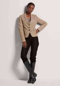 Beige op maat gemaakte blazer met drie knopen en twee voorkruistassen, gecombineerd met zwarte aansluitende broek en kniehoge zwarte leren laarzen.