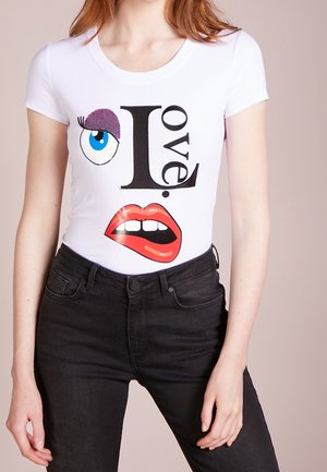 T-shirt blanc à manches courtes avec un design graphique représentant un œil, des lèvres, et le mot « amour » en noir, avec un accent violet brillant sur la paupière.