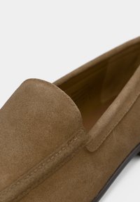 Gros plan sur une chaussure en daim marron à enfiler, avec un bout arrondi et des coutures visibles le long du bord supérieur et du côté.