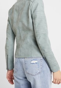 Ljusgrön lädermotorcykeljacka med texturerade ärmar, figursydd design och dragkedjeärmar; bärs med ljusblå slitna jeans.