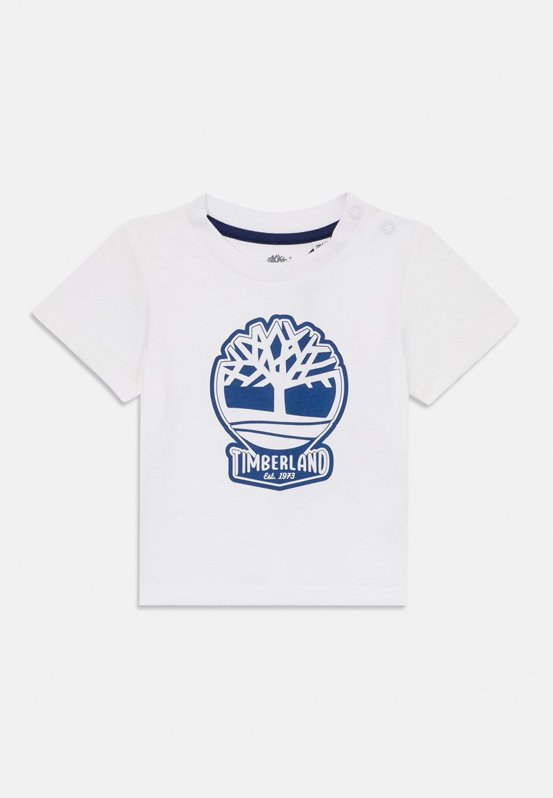 Camiseta de algodón blanca con mangas cortas, que presenta un logo de Timberland azul y un gráfico de árbol. Dos botones a presión en el hombro para facilitar el vestido.