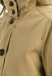 Khaki trenčkot s vysokým límcem, se čtyřmi tmavými knoflíky a hladkým, lehkým materiálem. Detailní šití podél švů.