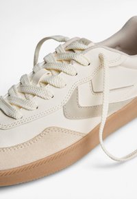 Baskets en cuir crème, avec des accents en daim marron clair, une semelle en caoutchouc texturée, et des lacets beige. Sur le côté, un motif discret gris clair.