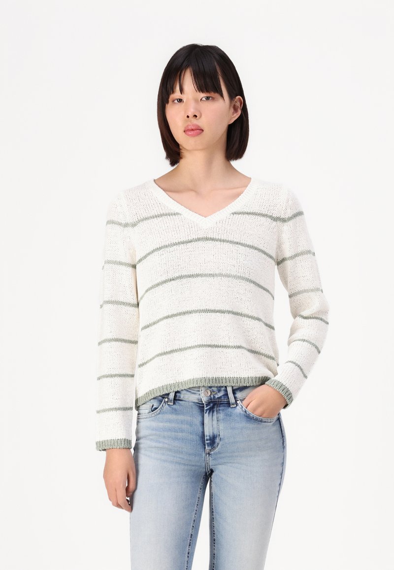 Hvit strikket genser med grønne striper, V-halsdesign, lange ermer og ribbestrikkede ermer. Kombinert med blå denimjeans.