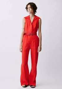 Combinaison rouge en lin avec un col, sans manches, jambes larges et évasées, et une ceinture texturée assortie. Chaussures noires à bout ouvert.