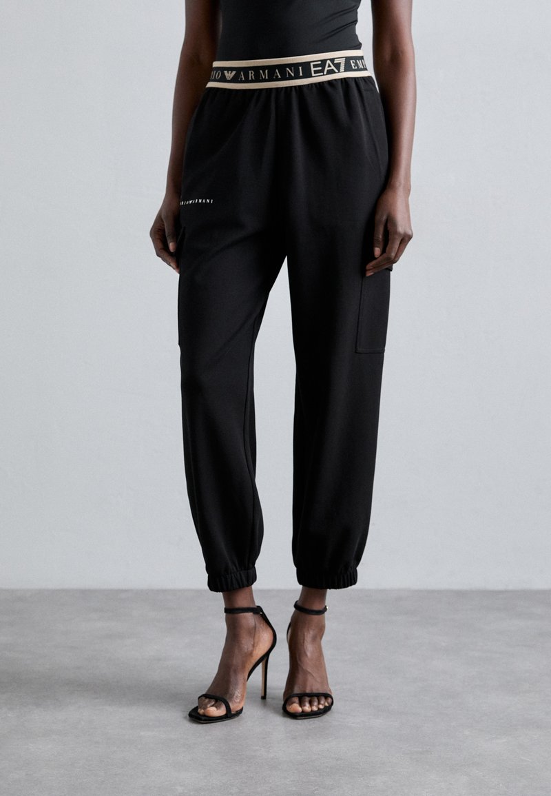 Melni džoginga bikses ar zelta logo jostasvietu, elastīgām piedurknēm un sānu kabatu; gluda auduma, ar brīvu fit.