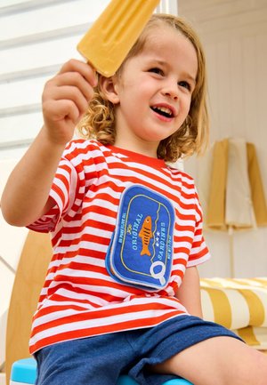 Nuori lapsi punavalkoraidallisessa paidassa, jossa on sardiinipurkin merkki, pitää oranssia mehujäätä ylhäällä hymyillen sisätiloissa raidallisen tyynyn lähellä.