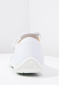Białe, błyszczące syntetyczne buty z rzepem, zaokrąglonym tyłem i teksturowaną gumową podeszwą. Prosty design, minimalne akcenty.