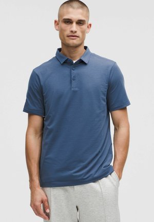 Jonge man met kort haar, gekleed in een blauwe polo met korte mouwen en lichtgrijze broek, staande met één hand in zijn zak.