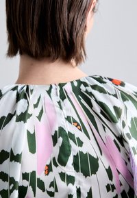 Bunt gemusterte Blumenbluse mit grünen, rosa und weißen Mustern, ausgestattet mit einem Reißverschluss hinten und gerafftem Stoff am Ausschnitt. Glatte Textur.