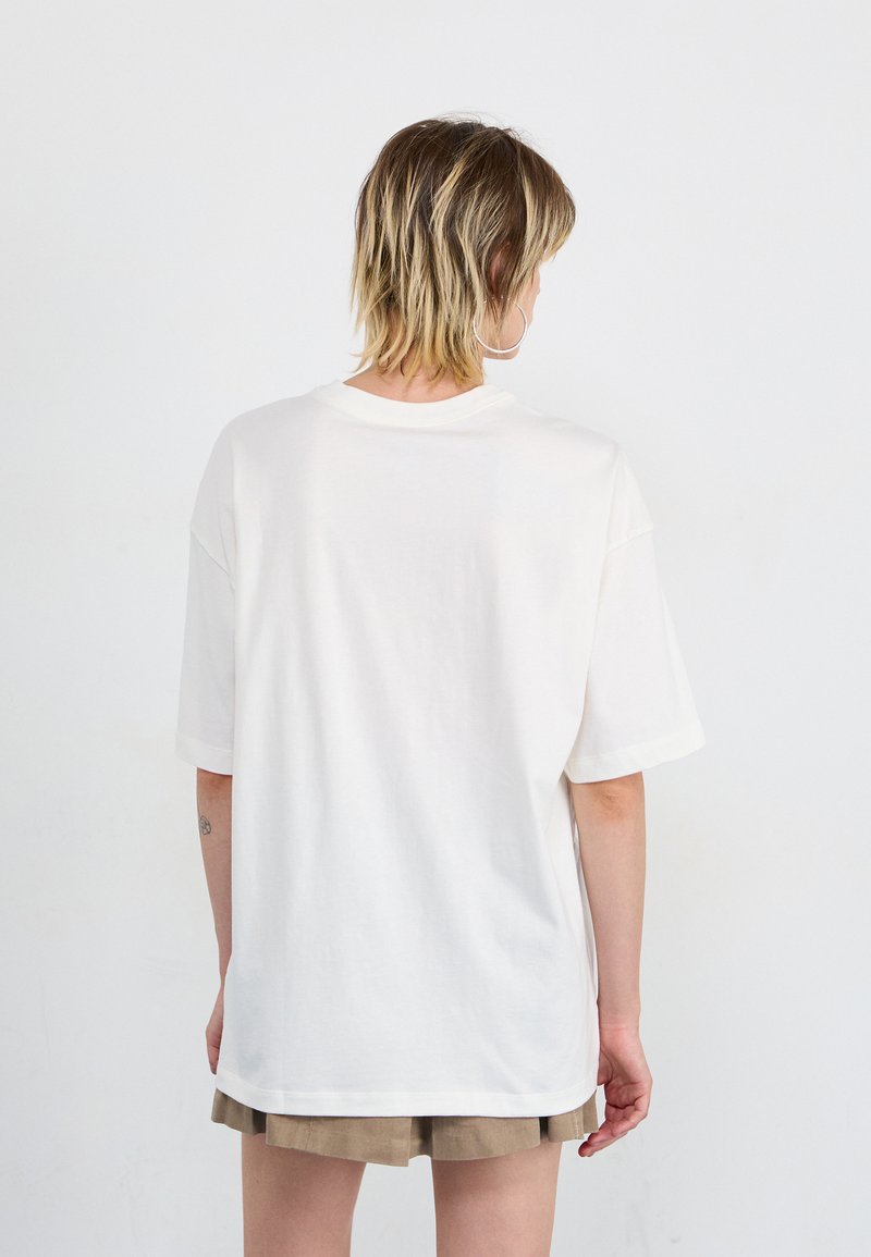 Übergroßes weißes Baumwoll-T-Shirt mit kurzen Ärmeln, Rundhalsausschnitt und geradem Saum, getragen über hell-beigen Shorts.