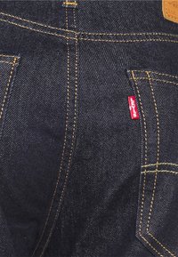 Jeans de ganga escura com costura visível, apresentando uma etiqueta vermelha da Levi's no bolso. Tecido durável e design padrão de cinco bolsos.