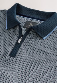 Tengerészkék galléros póló geometriai mintával, texturált gallérral és cipzáras záródással. Kék díszítésekkel és "NEXT REGULAR" feliratos címkével.