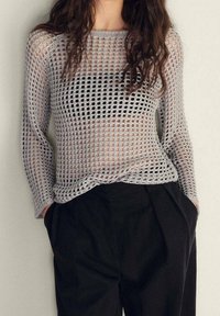 Femme portant un haut gris à manches longues en maille ouverte et ample par-dessus un soutien-gorge noir, associée à un pantalon large noir taille haute, les mains dans les poches.