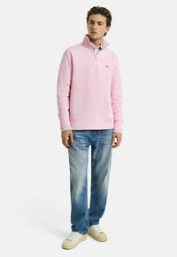 Pullover in pile rosa con colletto a mezza zip, abbinato a jeans azzurri chiari e scarpe da ginnastica bianche. Presenta un logo discreto sul petto.