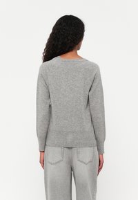 Sweater cinzento de gola redonda com mangas longas, bainha canelada e detalhe de gola em formato de escalope. Tecido de malha texturizado. Usado com jeans de cintura alta cinzentos claros.