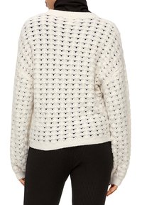 Weißer gestrickter Pullover mit strukturiertem Muster, locker geschnitten und mit geripptem Saum. Darüber getragen eine schwarze Ripp-Hose.