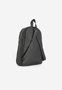 Zaino in pelle nera con forma arrotondata, cintura regolabile e chiusura con zip. Presenta una tasca laterale e una texture liscia.