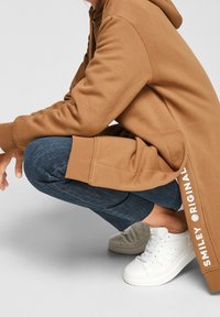 Sudadera marrón con la parte trasera larga, tejido texturizado, abertura lateral y texto "SMILEY ORIGINAL". Jeans azul oscuro y zapatillas blancas con perforaciones.