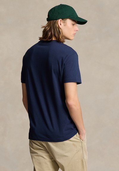 Young man wearing navy blue t-shirt, beige pants, and green baseball cap, standing with hands in pockets, facing sideways against plain background.