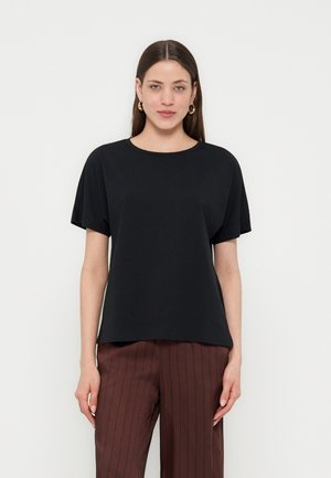 Kobieta o prostych brązowych włosach, nosząca złote kolczyki kółka, czarny krótkim rękawem top i brązowe spodnie w cienkie prążki, stojąca na białym tle.