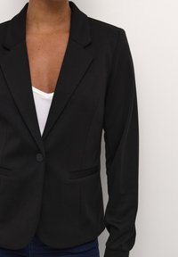 Czarna marynarka wykonana z teksturowanej tkaniny, z kołnierzem klapowym, z jednym guzikiem z przodu i dwoma kieszeniami, zestawiona z białą bluzką pod spodem.