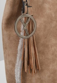 Brun taske i imiteret ruskind med en metallisk cirkulær vedhæng, der indeholder et 'X.' Vedhænget har læderkvaster og synlige syningsdetaljer.