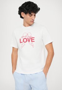 T-shirt de algodão branco com um gráfico rosa de dois querubins e a palavra "AMOR" em letras garrafais. Mangas curtas e decote redondo.
