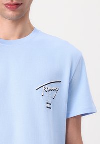 T-shirt en coton bleu clair à manches courtes. Présente un logo argent et noir sur la poitrine, ajoutant une touche élégante. Texture du tissu lisse.