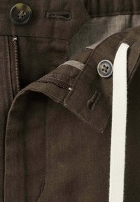 Gros plan d'une veste marron foncé montrant des boutons, des boutonnières, une fermeture éclair et un cordon blanc sur une texture de tissu.