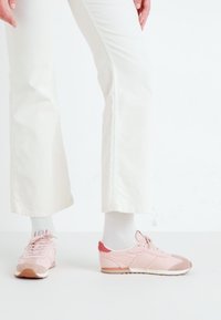Lys rosa joggesko laget av en kombinasjon av glatte og teksturerte materialer, med en rød detalj på hælen og en beige yttersåle.