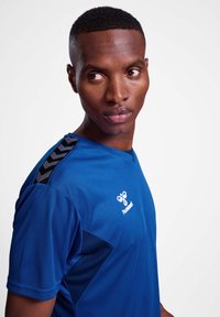 Blå atletisk skjorte lavet af glat stof, med V-udskæring og sorte detaljer langs skuldrene samt et logo på forsiden.