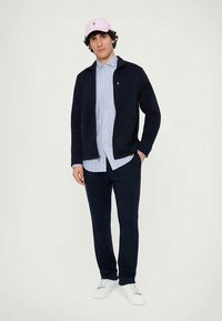 Marineblå glidelåsjakke, lyseblå og hvitstripet skjorte, marineblå bukser, hvite joggesko og en rosa caps med en logodetalj.