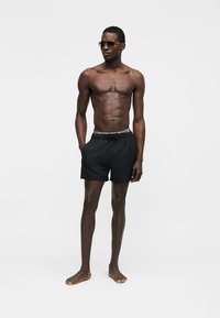 Särkset mees, kes kannab musti ujumispükse ja päikeseprille, seisab paljajalu, ühe käega püksitaskus, lakoonilise tausta ees.