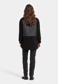 Musta fleece-takki, jossa on tikattu, matta nylonista valmistettu takapaneeli, pitkät hihat ja suora leikkaus. Yhdistetty kapeisiin mustiin farkkuihin ja mustiin jalkineisiin.