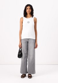 Camisola bege canelada com um gráfico de sol preto, combinada com calças largas cinzas e sapatos planos com padrão animal, segurando uma pequena bolsa preta.
