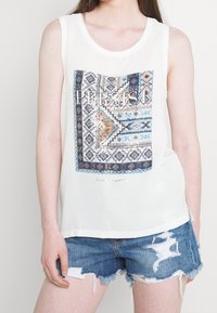 Biała bezrękawnikowa bluzka z niebieskim i pomarańczowym geometrycznym wzorem z napisem "PERSIST" oraz błyszczącym akcentem. Połączona z postarzanymi dżinsowymi szortami.