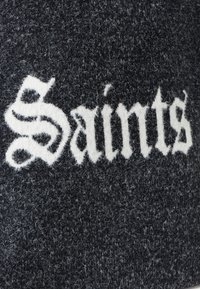 Czarna teksturowana tkanina zdobi śmiałe białe litery układające się w napis "Saints" w stylizowanej czcionce, co tworzy uderzający kontrast.
