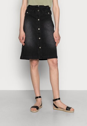 Frau trägt einen knielangen schwarzen Denimrock mit Knöpfen, Klapptaschen und schwarzen Espadrille-Sandalen mit Knöchelriemen.