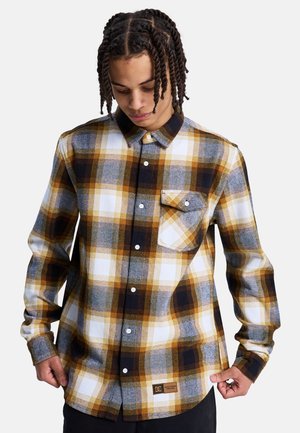 Koszula flanelowa w kratę z wzorem w kolorach żółtym, białym, szarym i czarnym, z kołnierzykiem na guziki, kieszenią na piersi oraz tkowaną metką przy dolnym brzegu.