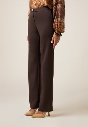 Žena stojící bokem, oblečená v tmavě hnědých rovních kalhotách s vysokým pasem, béžových špičatých podpatcích a vzorované dlouhém rukávu košili.