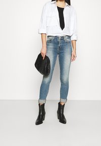 Ljusblå smala jeans med en blekt look, kombinerat med en svart topp och en lös vit skjorta. Svart quiltad handväska och svarta klackboots.