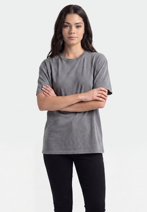 Ung kvinde med langt mørkt hår iført en enkel grå t-shirt og sorte bukser står med armene over kors mod en ensfarvet lys baggrund.