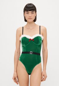 Bodysuit vert en velours avec des bordures en fausse fourrure blanche, un accent en ruban rouge, et une ceinture noire avec une boucle en strass. Bretelles noires réglables.