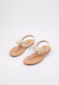 Sandalias planas beige con un diseño de tira trenzada y un adorno floral hecho de pequeñas perlas en la parte delantera. Suela de goma plana.