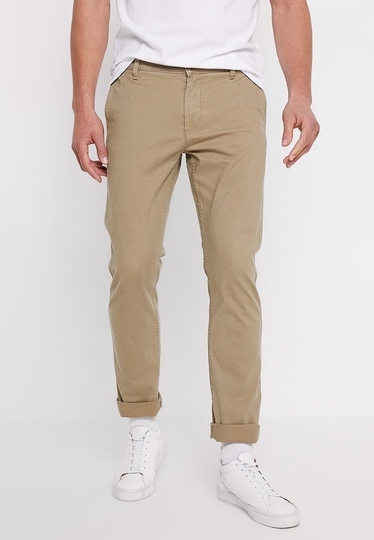 Chinos beiges avec une coupe slim, ourlets retroussés et poches avant, fabriqués à partir d'un tissu lisse. Portés avec un T-shirt blanc et des baskets blanches.