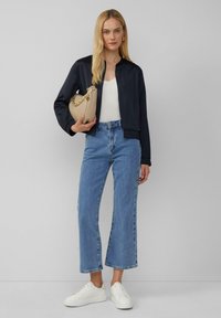 Veste de bombardier navy, débardeur blanc, jean large bleu clair et baskets blanches. Tenant un sac beige avec une accentuation en chaîne. Design simple.